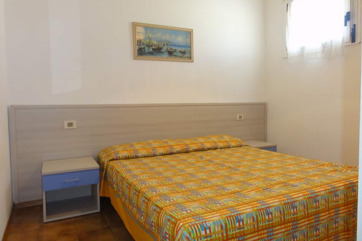 Un dormitorio con cama, mesita de noche, una pequeña mesa y un cuadro en la pared.