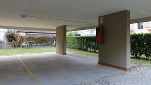 A covered parking area with a fire alarm box on the wall, a yellow line marking, and a building with a tiled roof in the background. - Condominio Isola Clara (San Michele al Tagliamento)