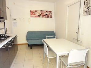 Een compacte keuken met een fornuis en kastjes, een blauwe bank, een witte eettafel met stoelen, en een aan de muur hangend bloemenportret.