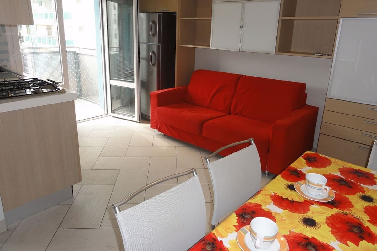 A modern kitchen with a red sofa, dining table with floral tablecloth, and a refrigerator.