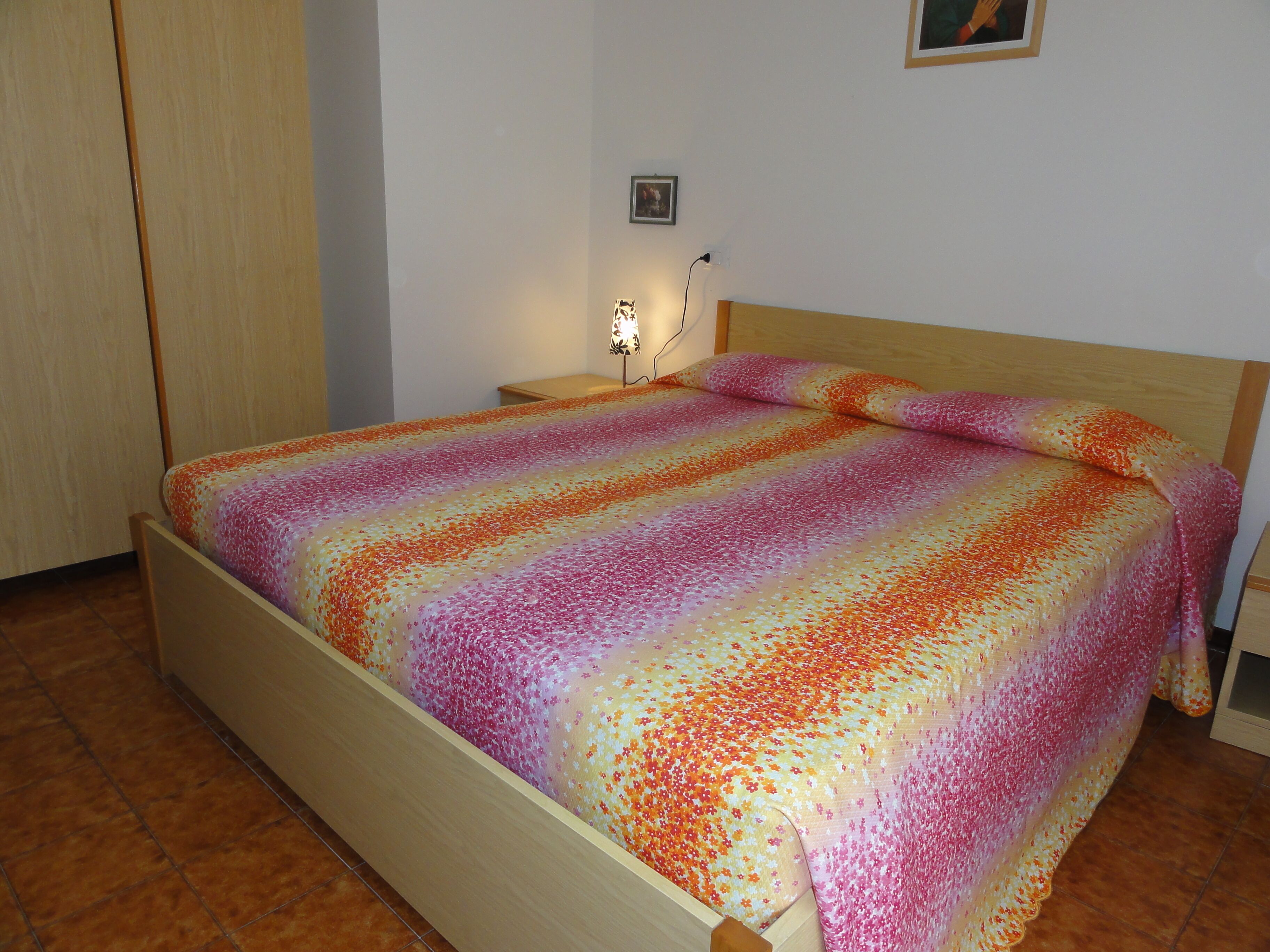 A bed with a floral-patterned bedspread, a wooden headboard, a nightstand with a lamp, and a wall-mounted picture.