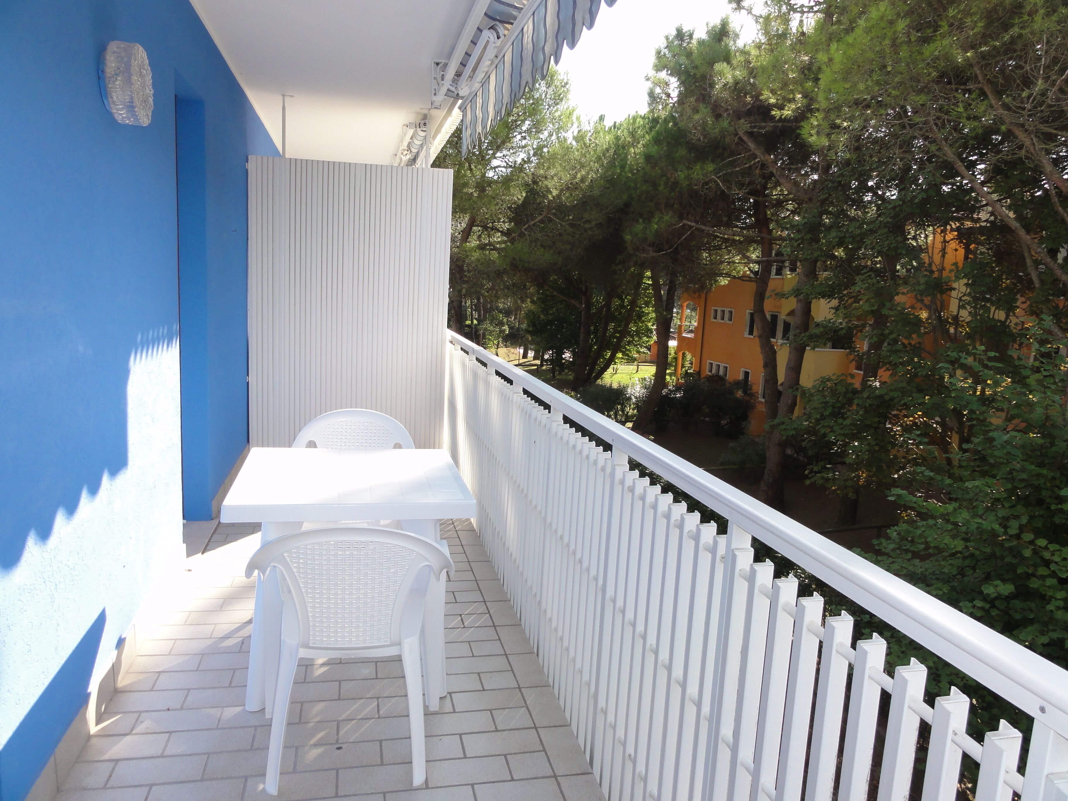 A balcony with a white table and chairs, a white picket fence, and a view of trees and buildings.