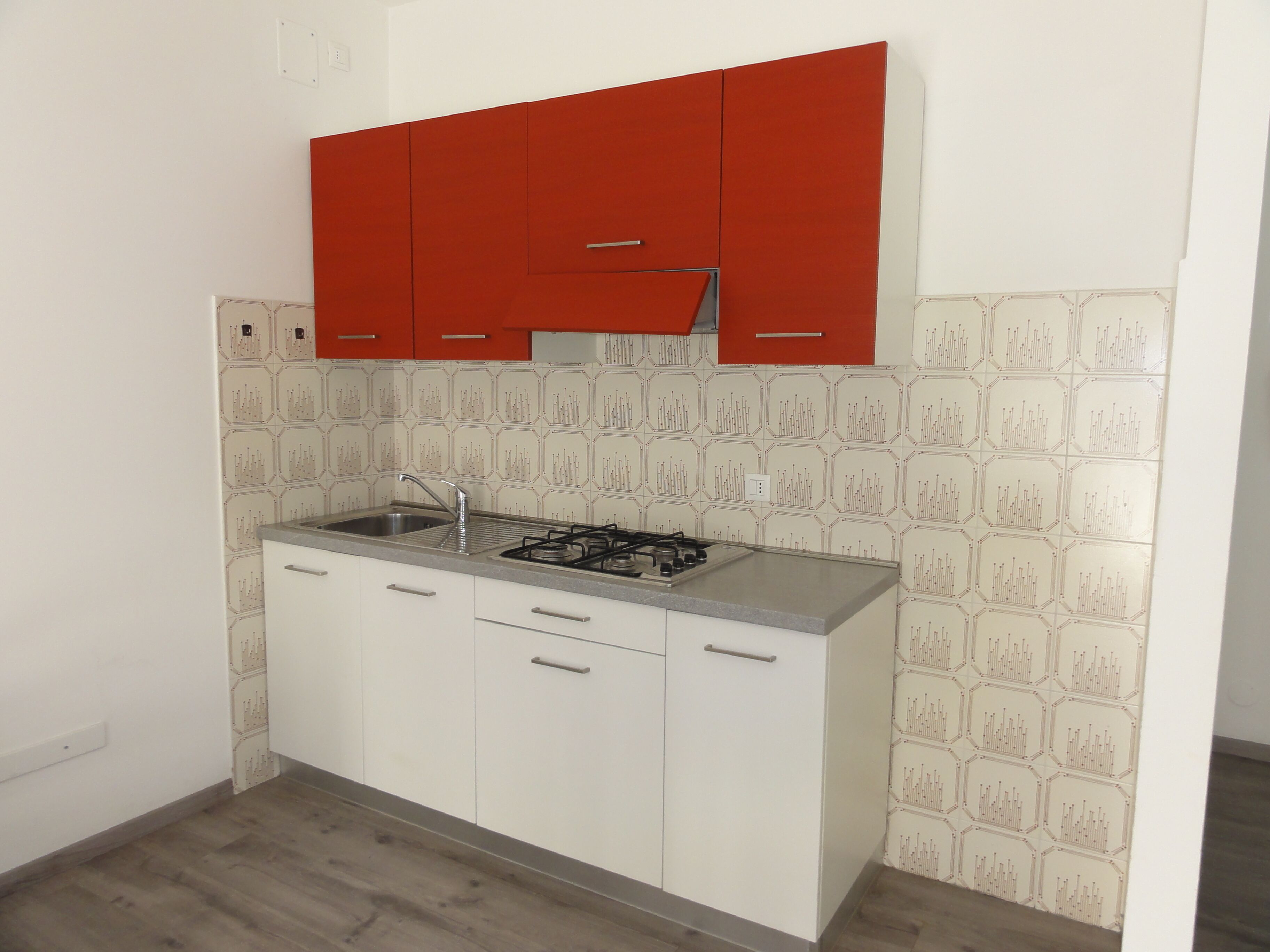 Une cuisine avec des meubles blancs, un meuble supérieur rouge, une cuisinière à gaz et un crédence à motifs.