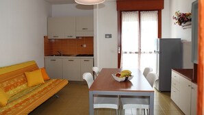 A compact kitchen with white cabinets, a dining table with chairs, and a sofa with orange cushions. - Condominio Simma 2 (San Michele al Tagliamento)