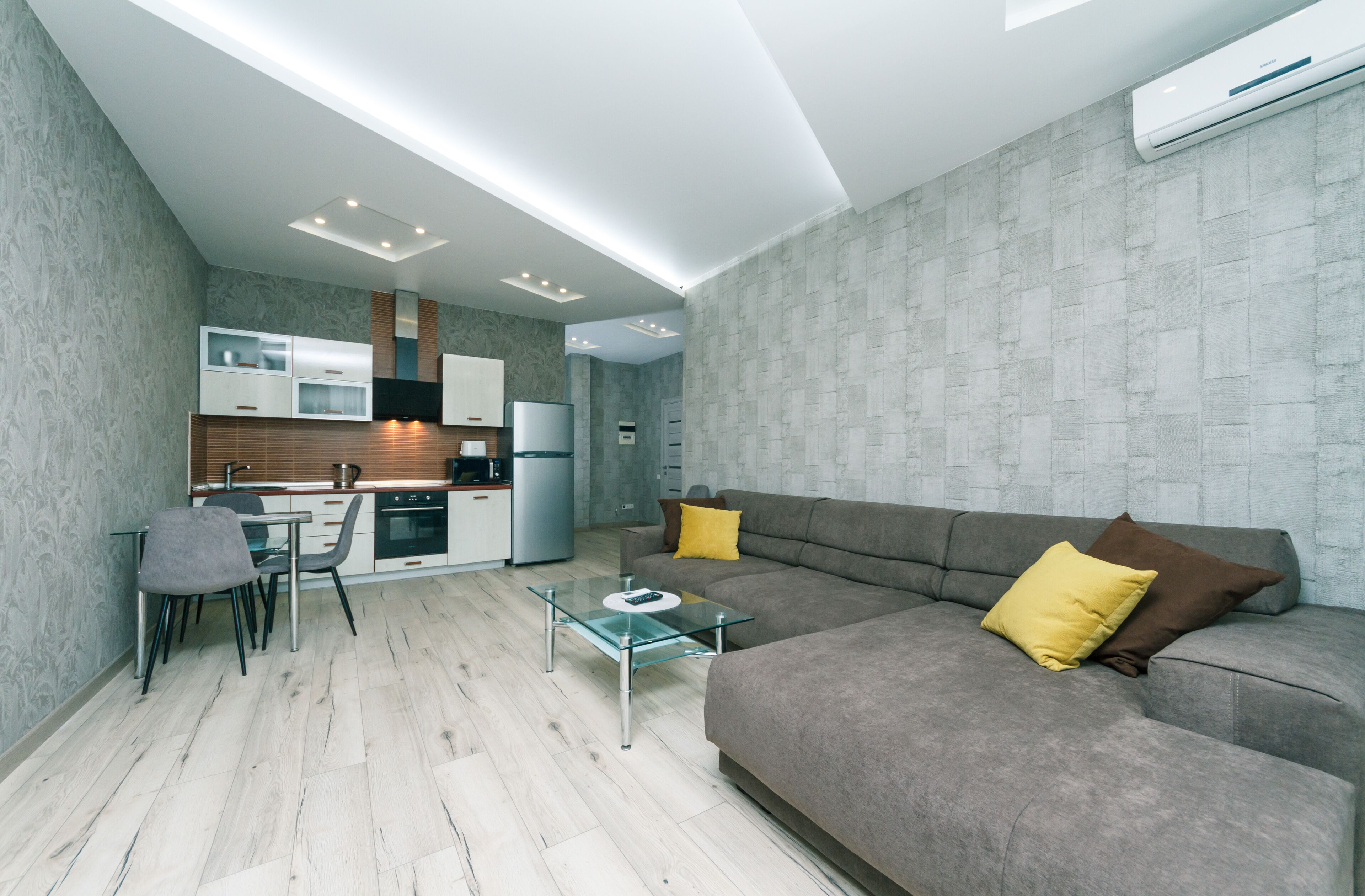 Uma sala de estar moderna com uma mesa de centro de vidro, sofá cinzento e um espaço de refeições com cozinha ao fundo.