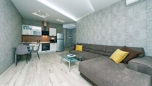 Ein modernes Wohnzimmer mit Glastisch, grauem Sofa und einem Essbereich, im Hintergrund ist eine Küche zu sehen.