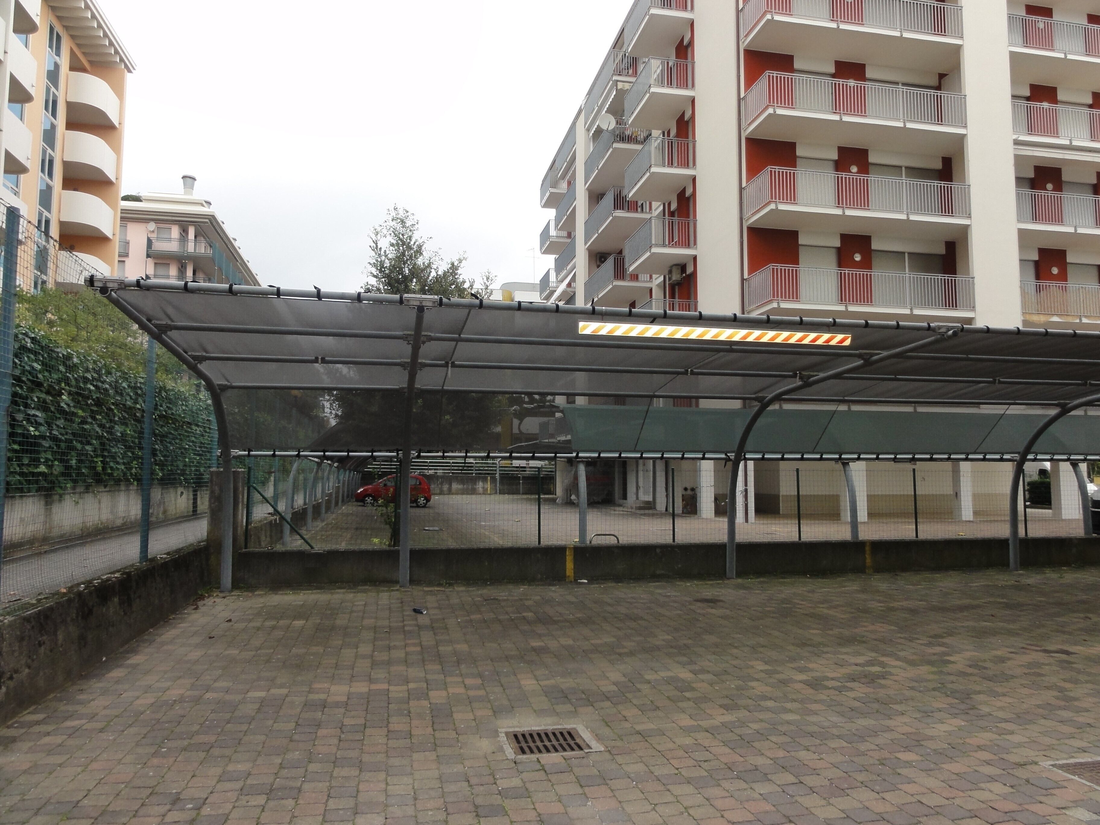 Ein überdachter Parkbereich mit Metalldach, einem gepflasterten Weg und einem roten Auto darin.