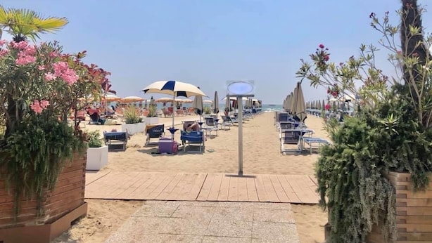 A beach with rows of sun loungers and umbrellas, a wooden walkway leading to the beach, and potted plants on either side. - Condominio Storione (San Michele al Tagliamento)