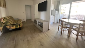 A living room with a sofa, a TV mounted on the wall, a dining table with chairs, and a kitchen area with a refrigerator and microwave. - Condominio Venere (San Michele al Tagliamento)
