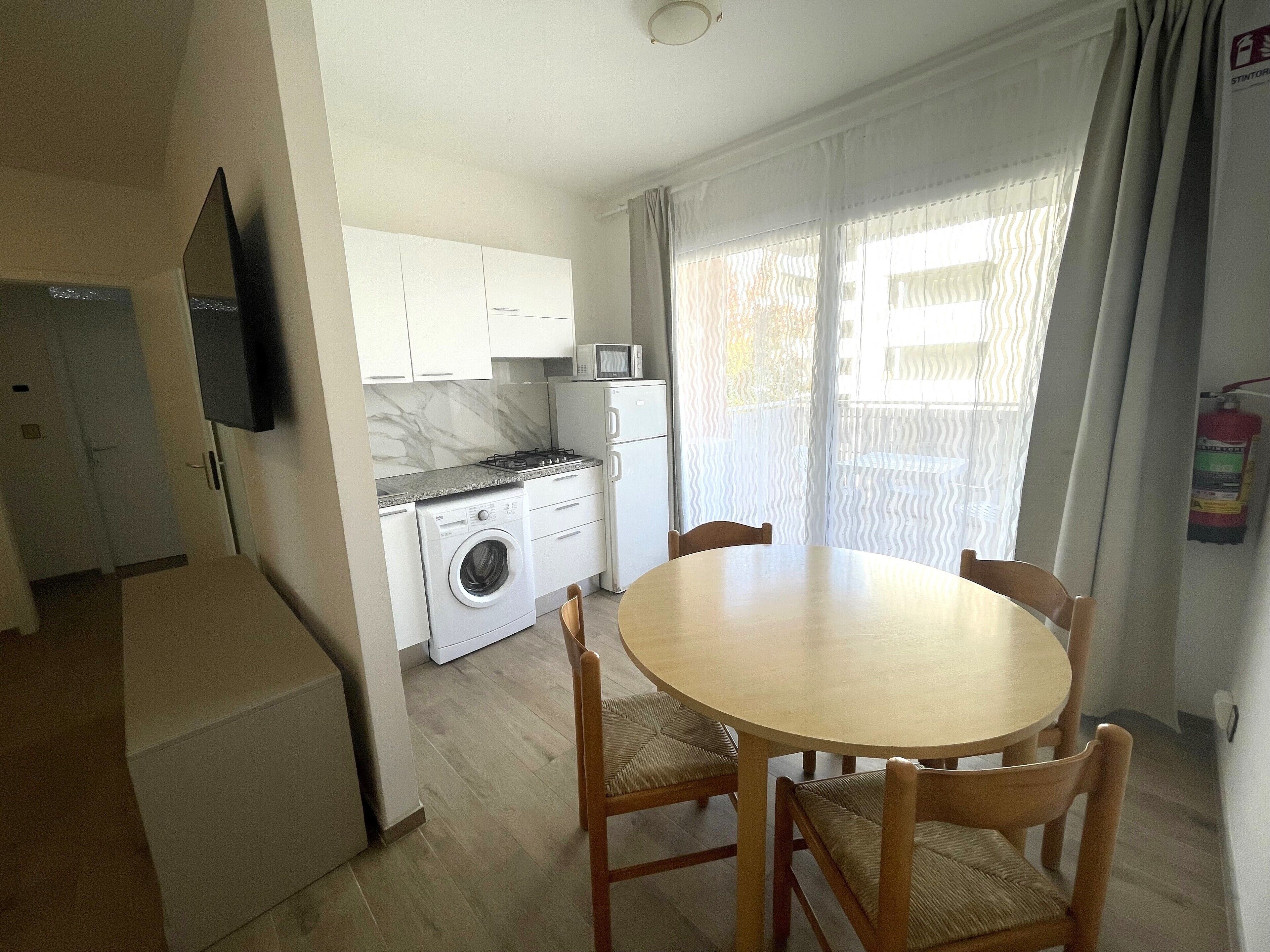 A compact kitchen with a washing machine, a microwave, and a stove. There is a dining table with chairs and a television mounted on the wall.