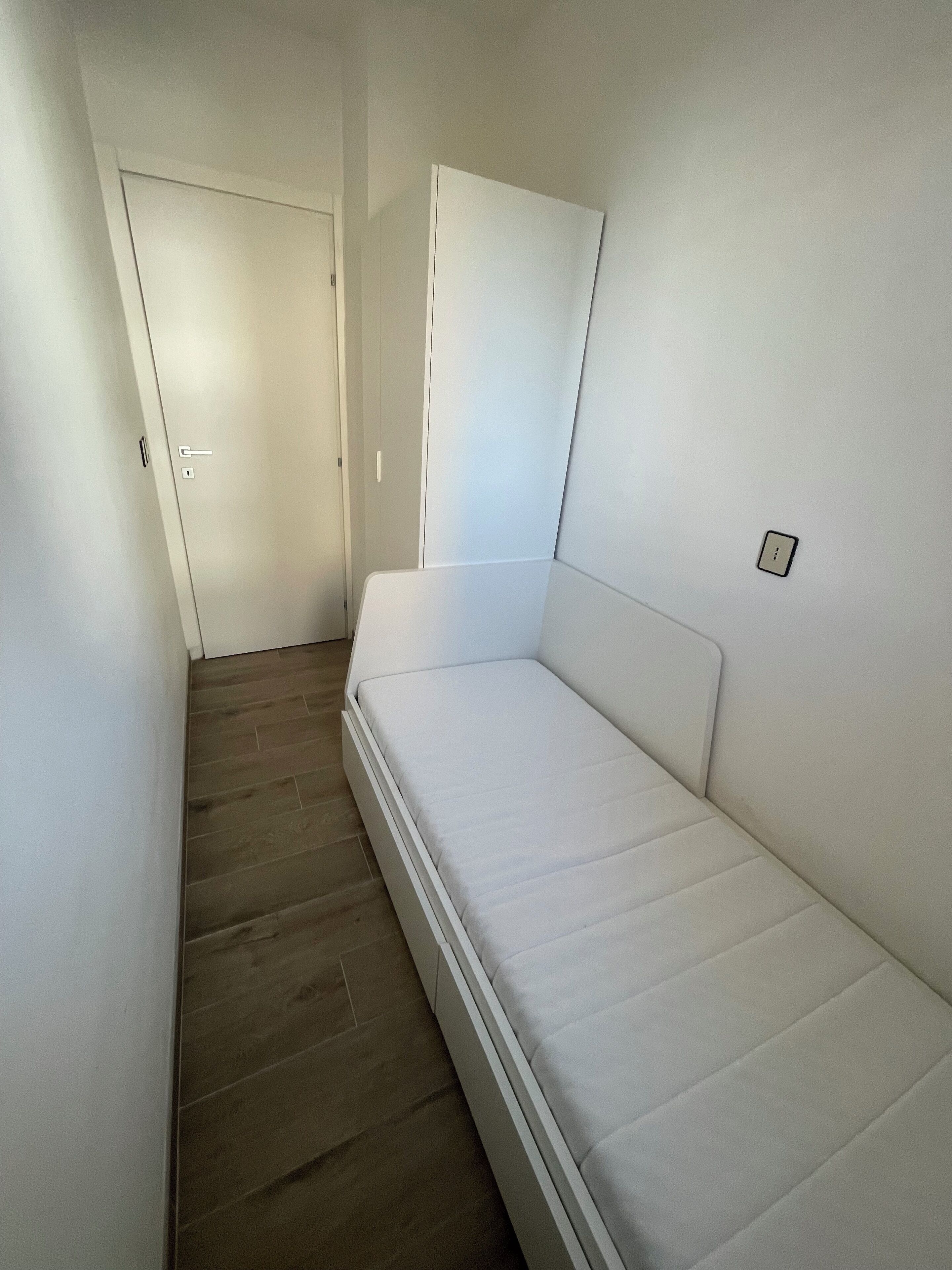 A narrow room with a single bed, a door, and a light switch on the wall.