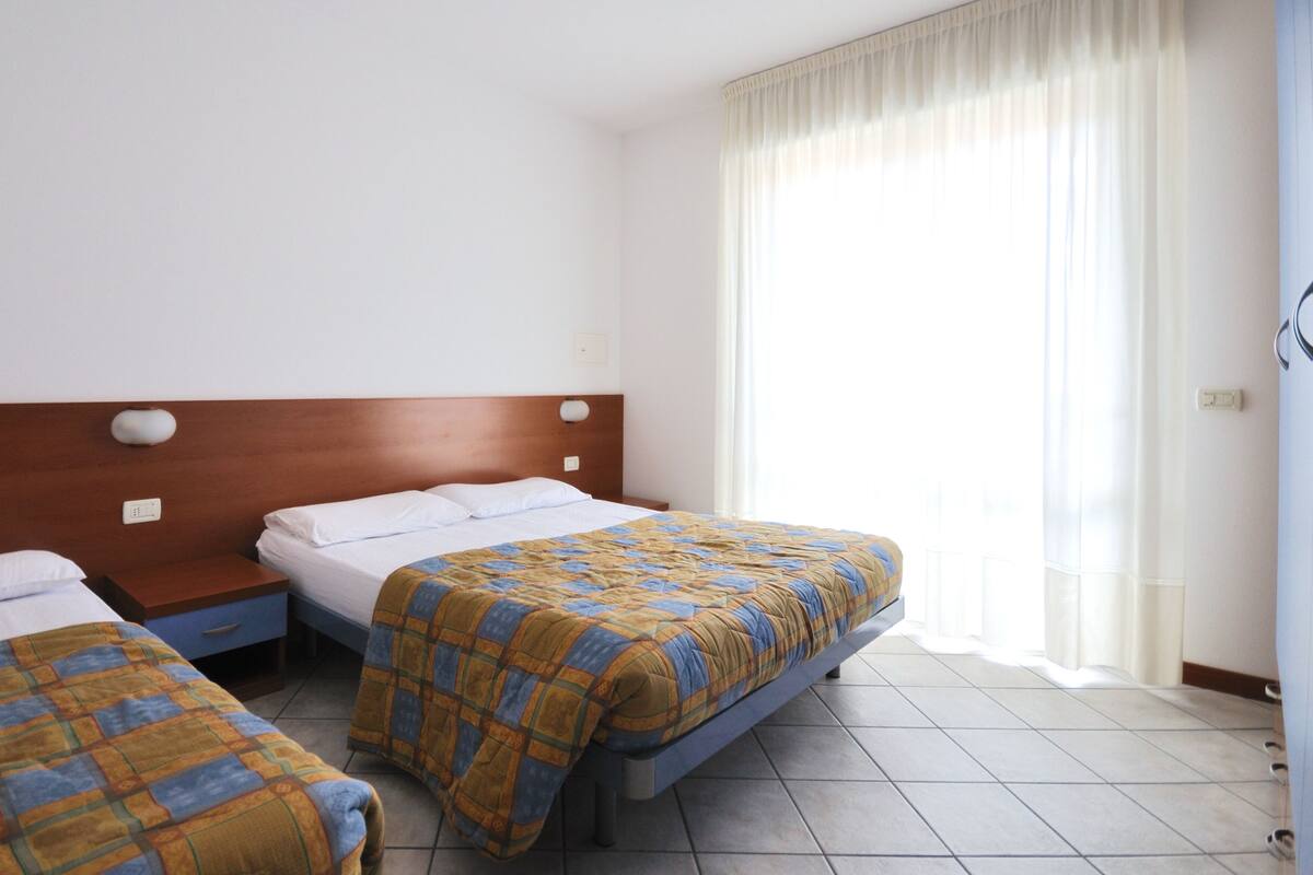 A hotel room with two beds, a wooden headboard, a window with curtains, and a tiled floor.