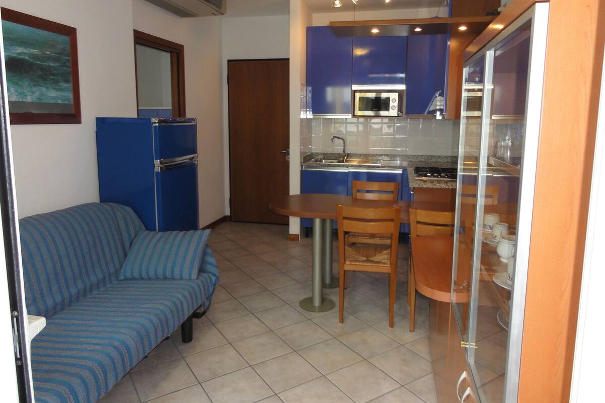 A compact kitchen with blue cabinets, a microwave, and a refrigerator. There is a dining table with two chairs and a sofa in the adjacent living area.