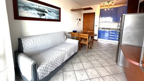 A living area with a sofa, a dining table with chairs, a kitchen with a refrigerator, and a door leading to a balcony. - Residence Eurostar (San Michele al Tagliamento)