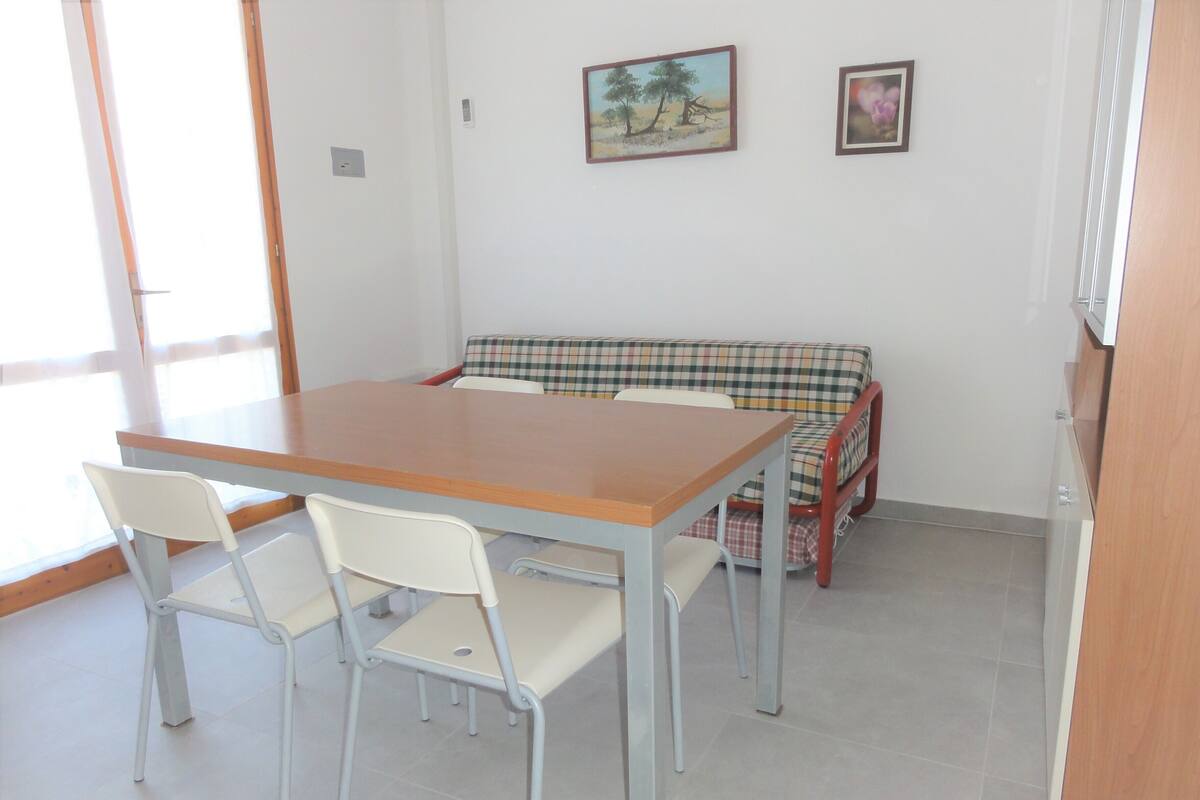 A dining area with a wooden table and two white chairs, a plaid sofa, and two framed pictures on the wall.