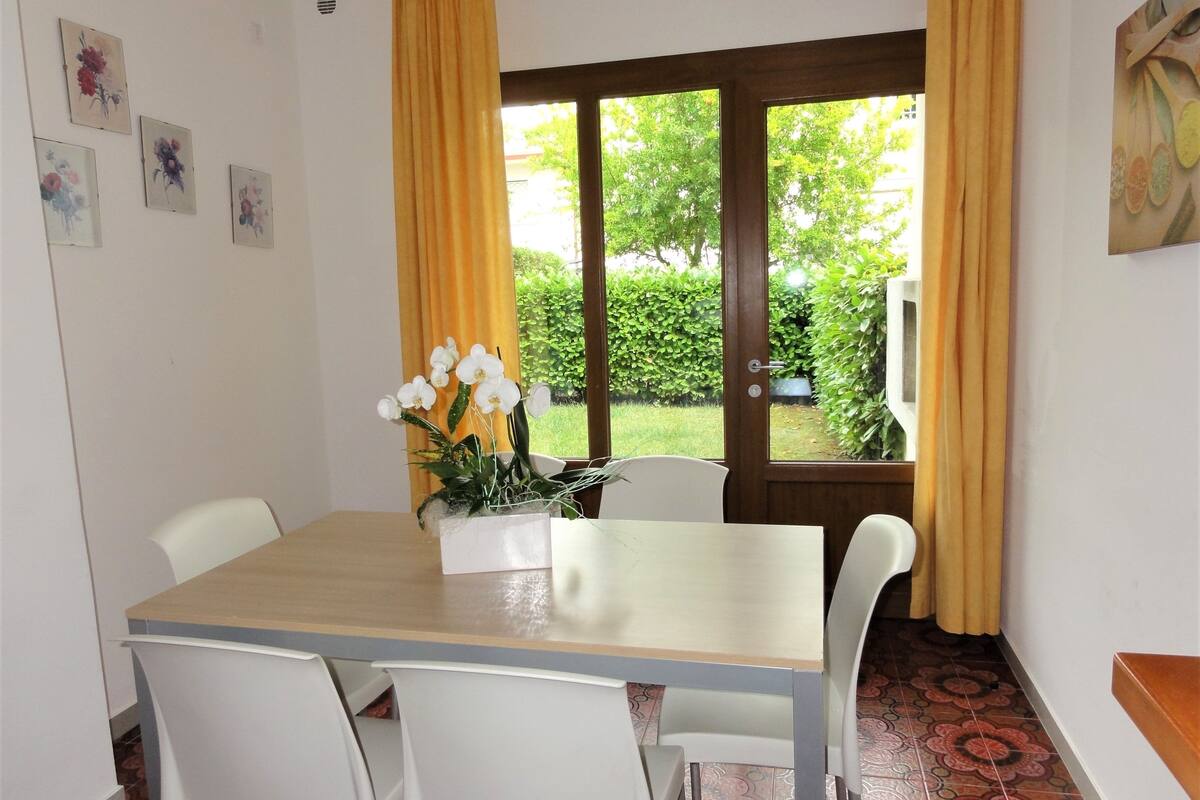 A dining area with a wooden table and white chairs, a vase of white flowers, and a window with yellow curtains.