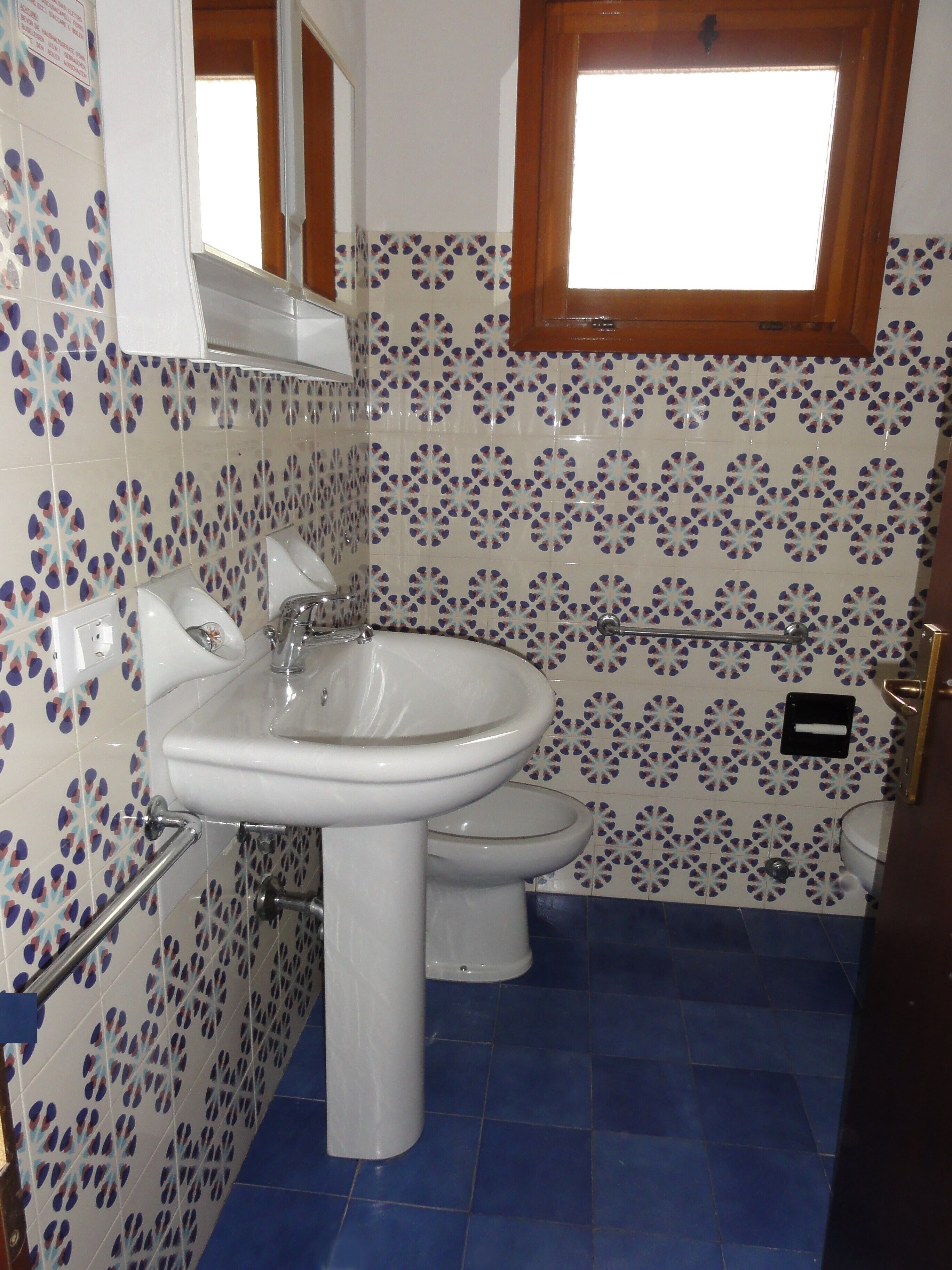 Une salle de bain avec un lavabo blanc, des toilettes et un bidet. Les murs sont recouverts de carreaux aux motifs bleus et blancs.