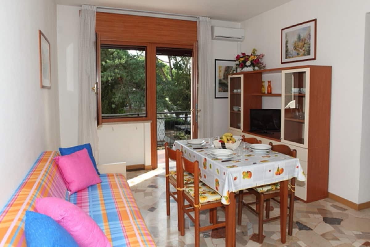Uma sala de jantar com uma mesa posta para quatro pessoas, um sofá com almofadas coloridas e um armário com portas de vidro.