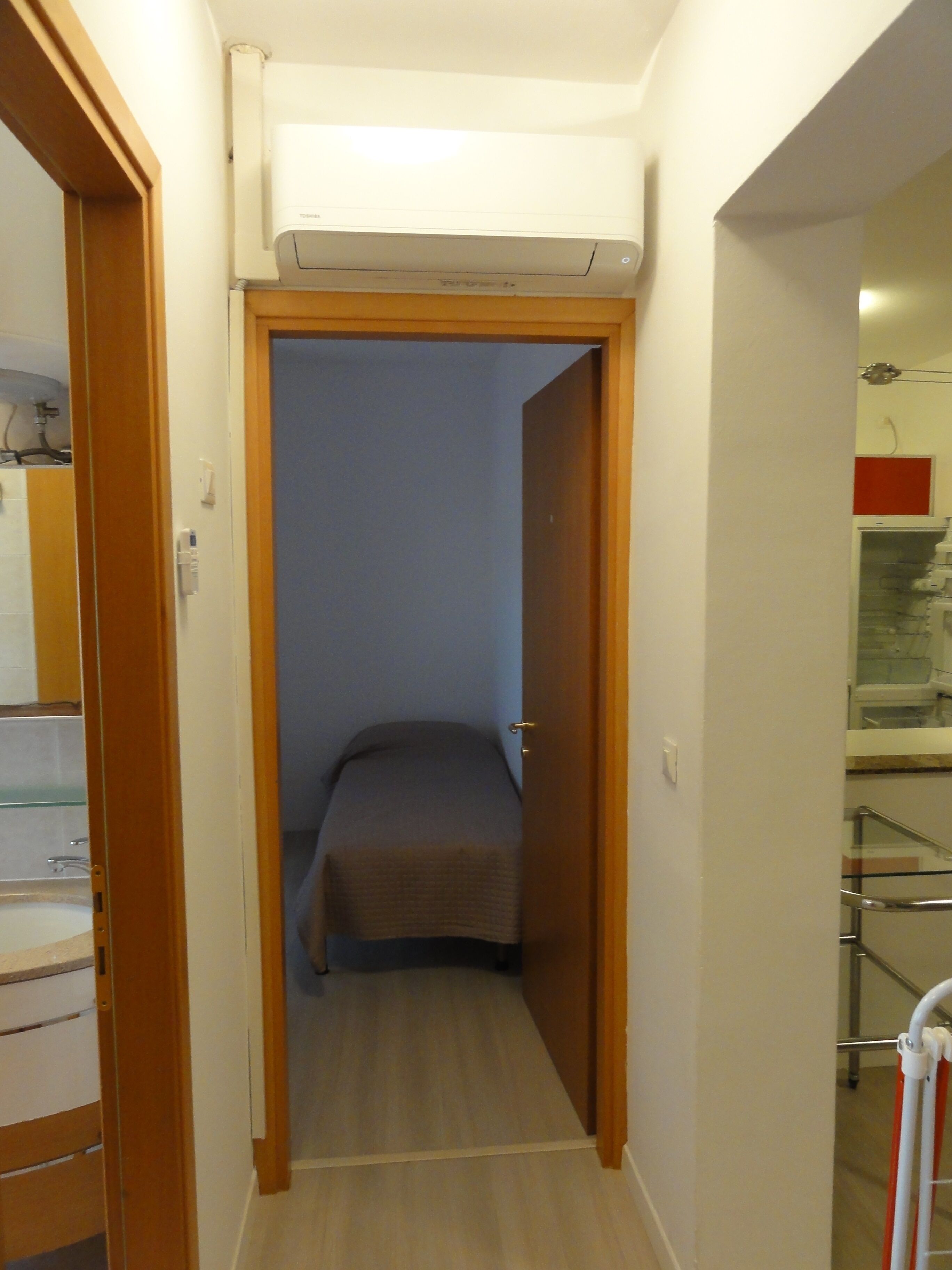 Um corredor estreito com uma cama no final, um aparelho de ar condicionado branco acima e um banheiro com chuveiro de vidro à direita.