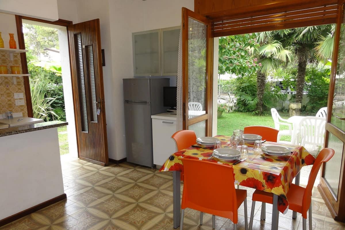 A dining area with a table set for four, surrounded by orange chairs, a refrigerator, and a television in the background.