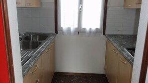 A compact kitchen with two sinks, wooden cabinets, and a window with white curtains. - Villa Letizia (San Michele al Tagliamento)
