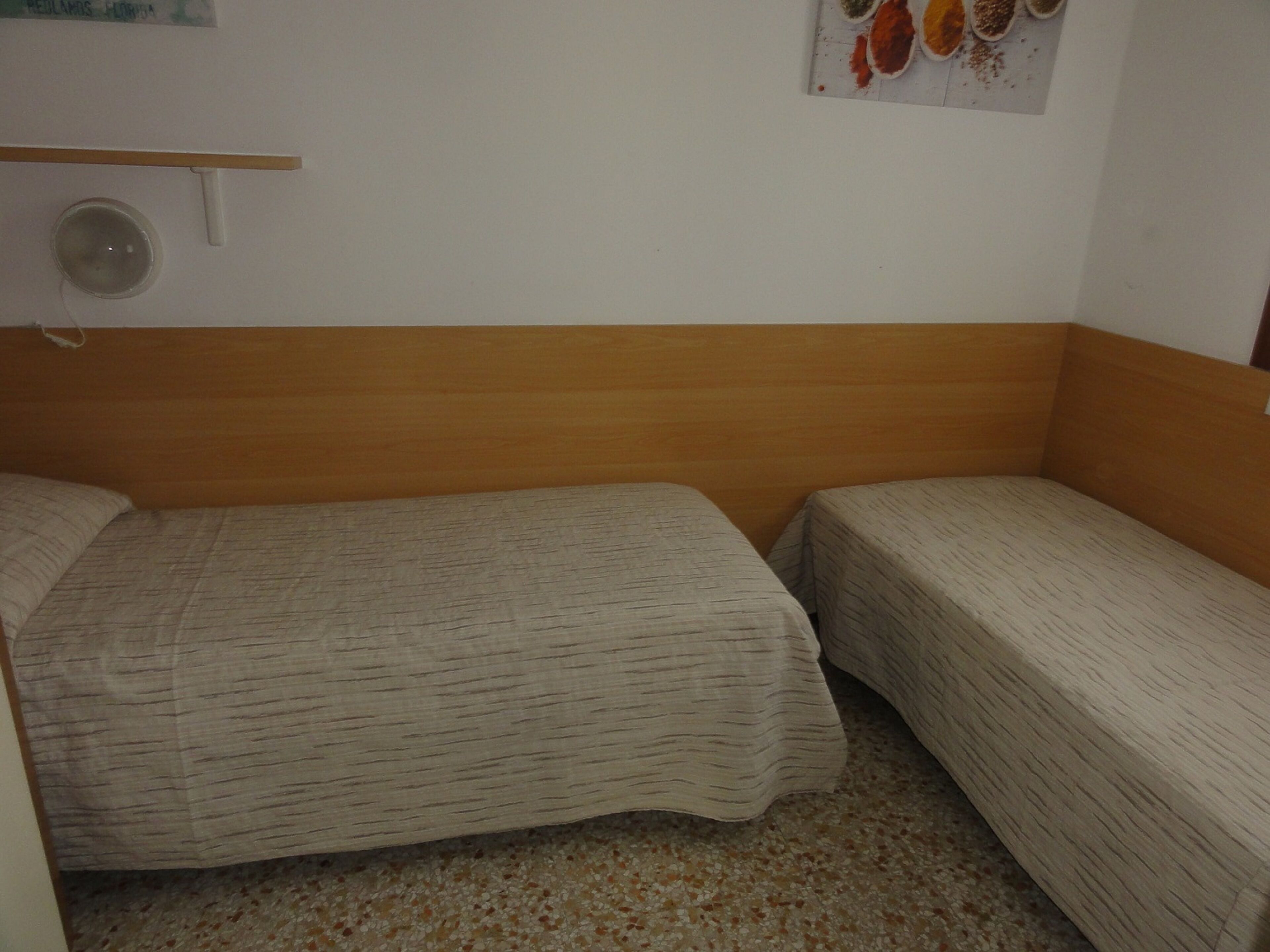 Ein kleines Hotelzimmer mit einem Bett, zwei Einzelbetten, einem hölzernen Kopfteil, einer kleinen Ablagefläche und einer Wandleuchte.