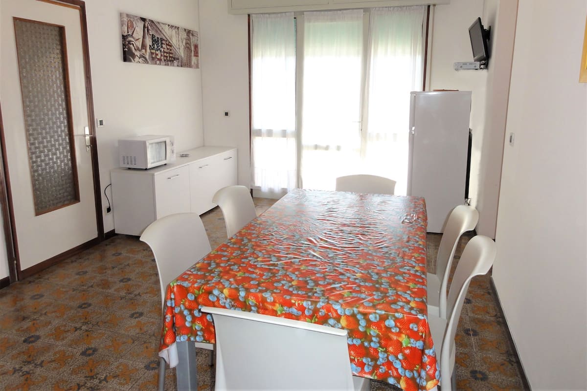 A compact kitchen with white cabinets, a microwave, and a refrigerator. There is a dining table with chairs, and a window with curtains.
