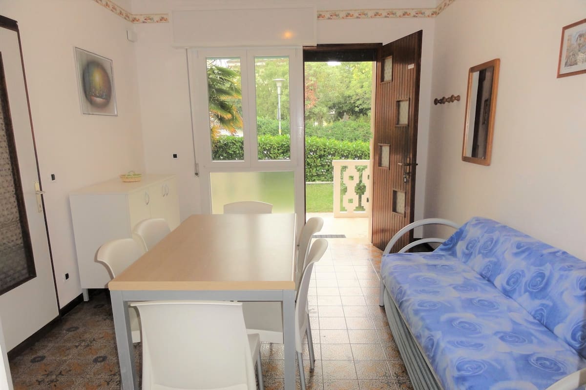 A room with a dining table, chairs, a sofa, and a view of a garden through a door.