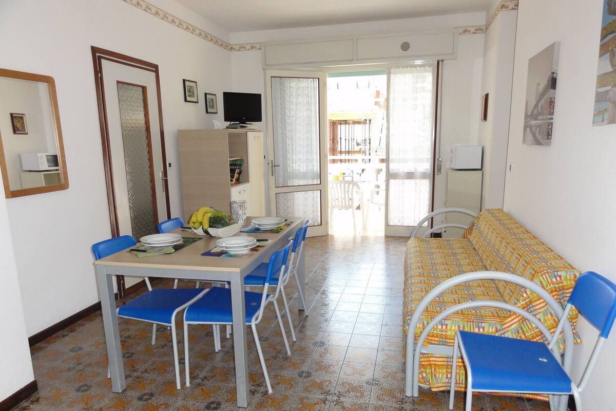 A dining area with a table set for four, blue chairs, a sofa with patterned cushions, and a television on a cabinet.