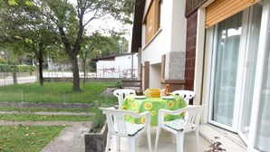 Um pátio externo com uma mesa posta para quatro pessoas, rodeado por cadeiras brancas, e uma grande janela com vista para a rua.