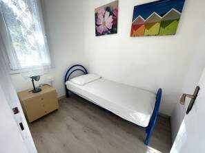 A small, single-bed room with a wooden floor, a bedside table with a lamp, and two wall-mounted paintings.