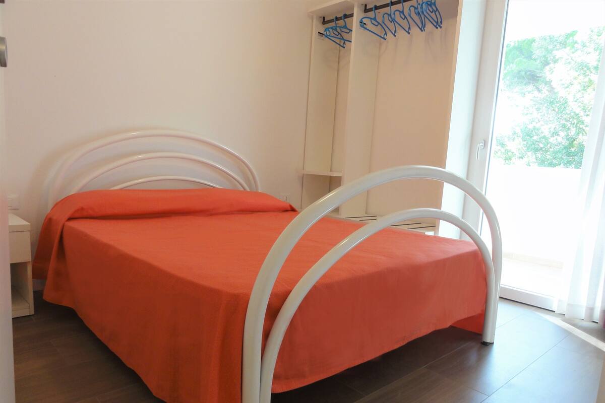 Un lit avec un cadre en métal blanc et une couvre-lit orange, dans une pièce au plancher en bois et dotée d’une fenêtre avec des rideaux.