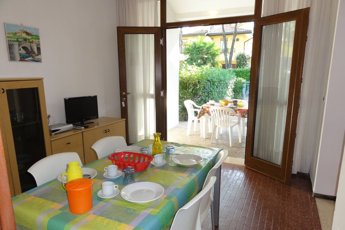 A dining area with a table set for four, a television on a cabinet, and a view of an outdoor patio with a table and chairs.