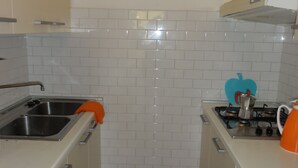 Una cucina compatta con mobili bianchi, un lavello in acciaio inossidabile, un piano cottura a gas e un dispenser di sapone a forma di mela blu.