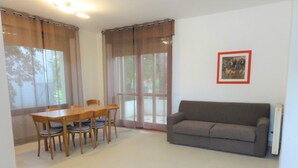 Ein Wohnzimmer mit Esstisch und Stühlen, einer Couch und einem großen Fenster mit Vorhängen.