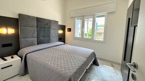 Un dormitorio moderno con una cama grande, una mesita de noche, una lámpara de pared y una ventana que ofrece vistas a edificios.