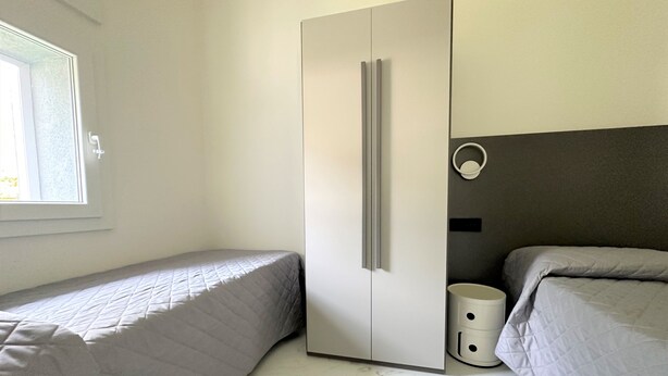 A bedroom with two beds, a wardrobe, a small trash bin, and a window. - Villa Wanda (San Michele al Tagliamento)