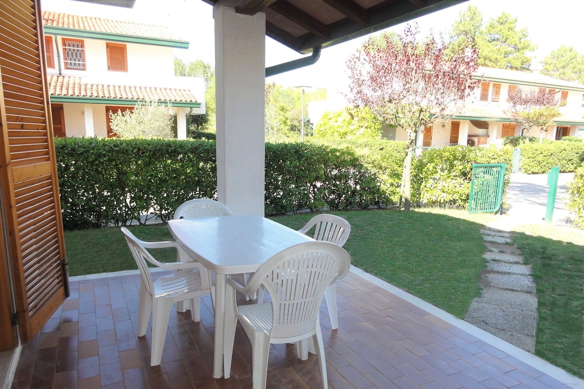 A covered patio with a white table and chairs, surrounded by a brick floor and a green hedge.