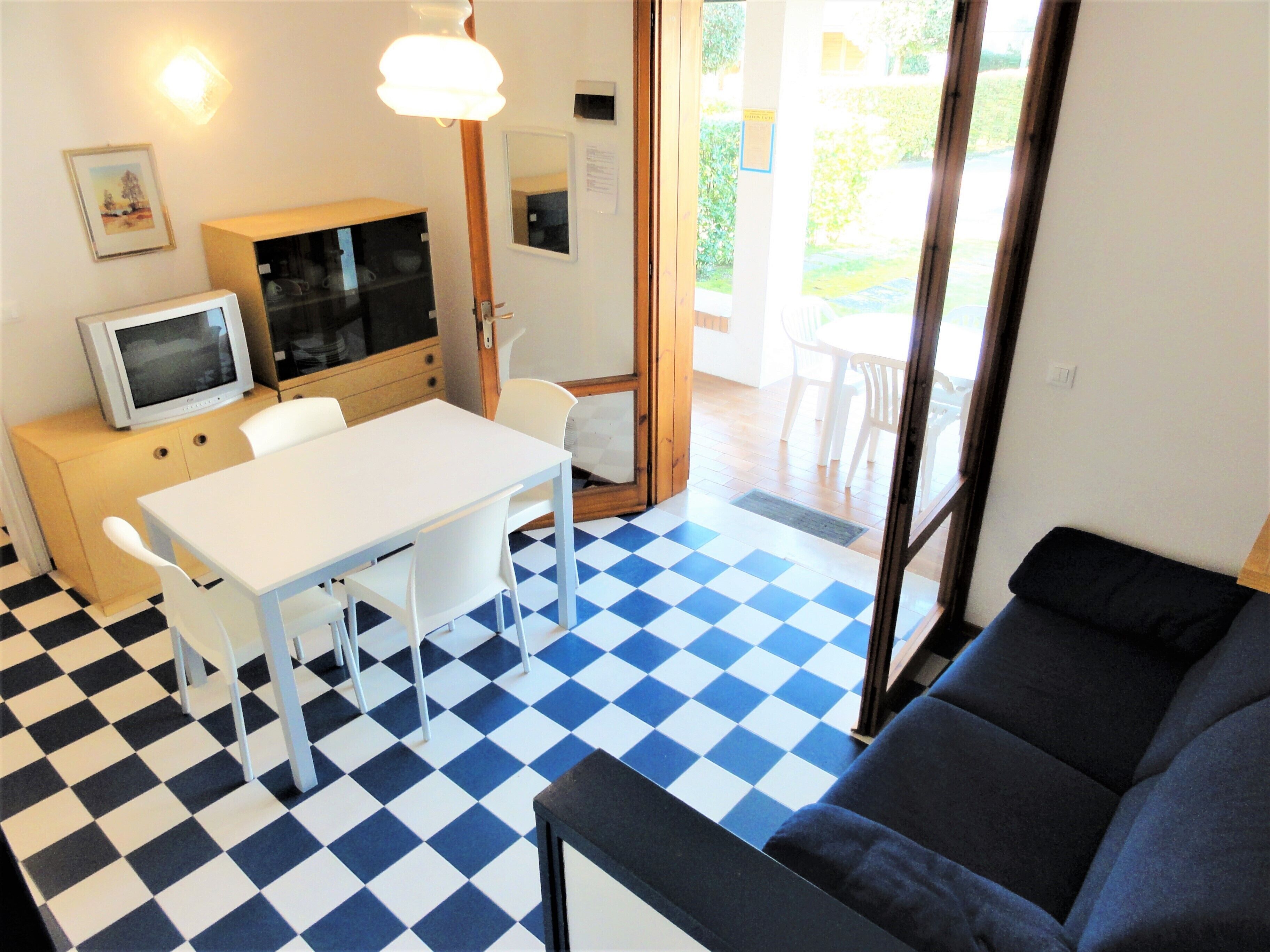 A room with a checkered floor, a white table with four chairs, a black sofa, a television on a wooden cabinet, and a door leading outside.