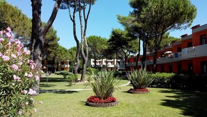 A courtyard with neatly trimmed grass, several trees, and a building with red and white exterior walls. - Villaggio White Star (San Michele al Tagliamento)