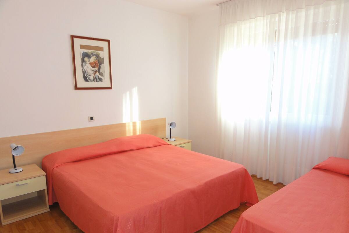 Habitación de hotel con dos camas, una mesita de noche con lámpara, un cuadro en la pared y una ventana con cortinas traslúcidas.