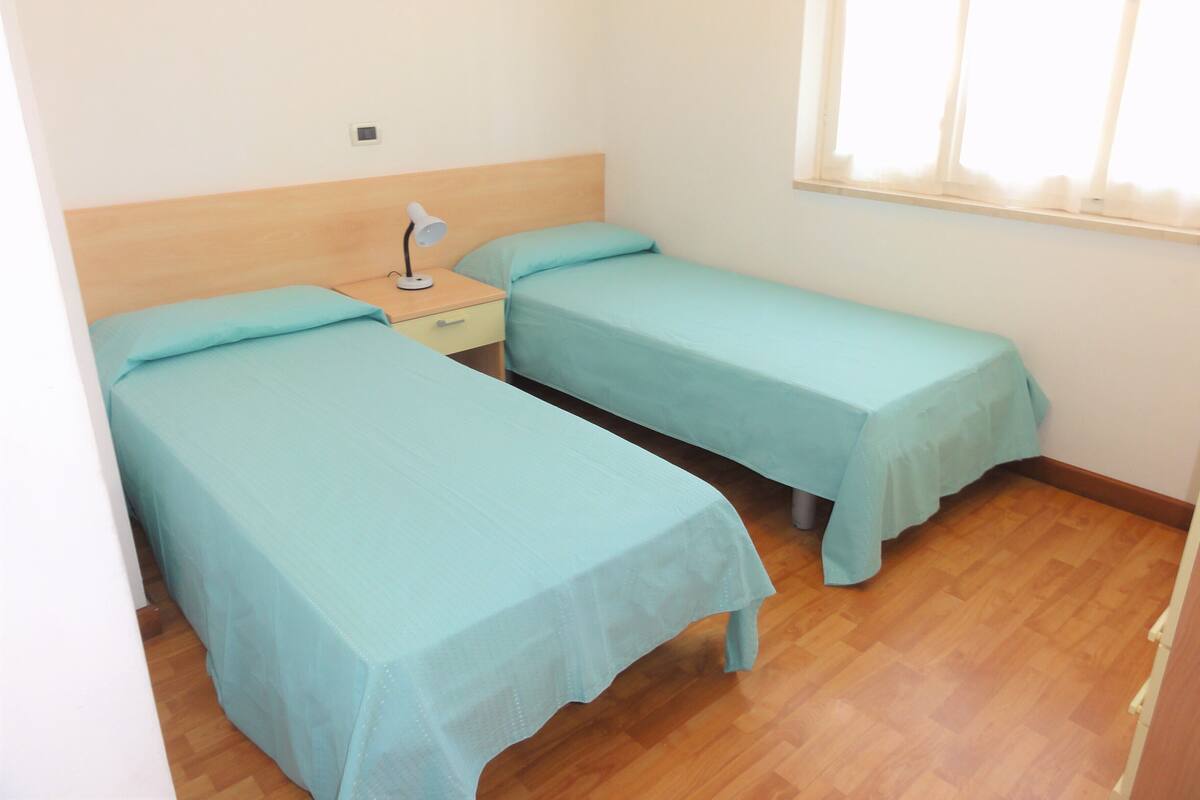 Una habitación pequeña y limpia con dos camas individuales, una mesita de noche con lámpara y una ventana con cortinas.