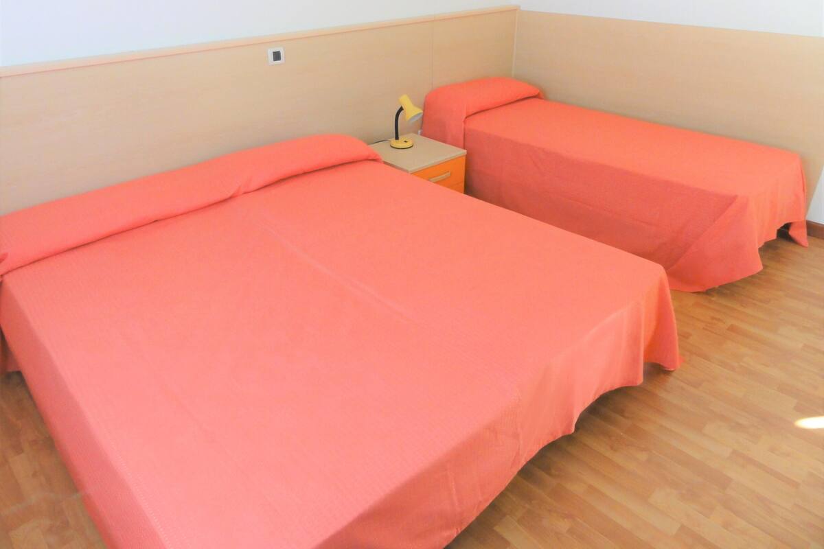 Una habitación con dos camas individuales, cada una con un edredón naranja a juego, una mesita de noche pequeña con una lámpara y suelo de madera.