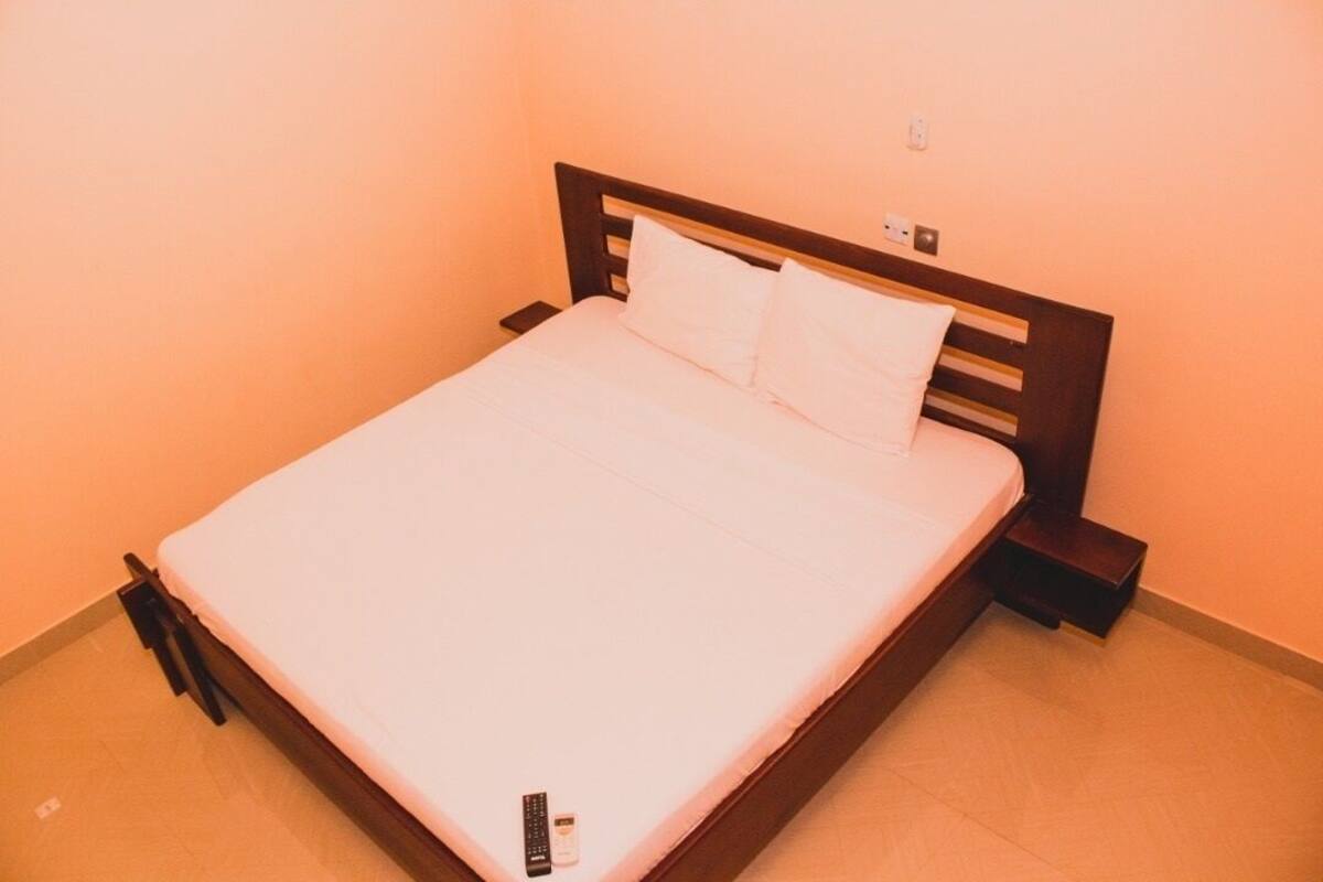 A single bed with a wooden headboard, a white mattress, and two pillows. There is a remote control and a small bottle on the bed.