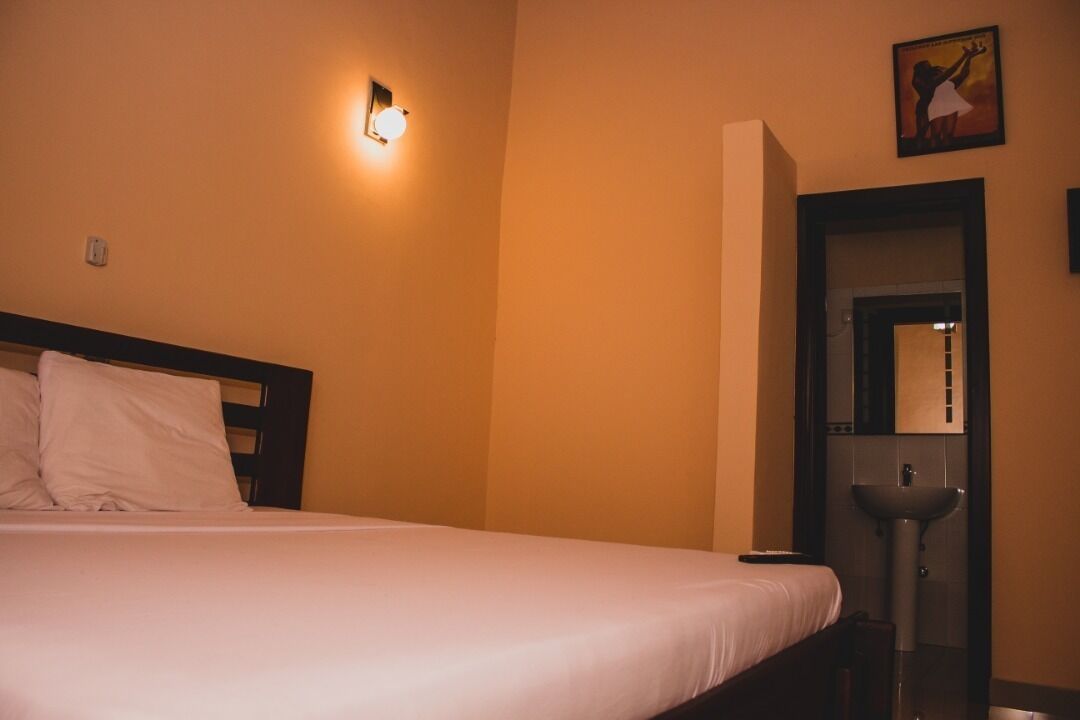 A hotel room with a bed, a nightstand, a wall-mounted light, a door, and a sink.