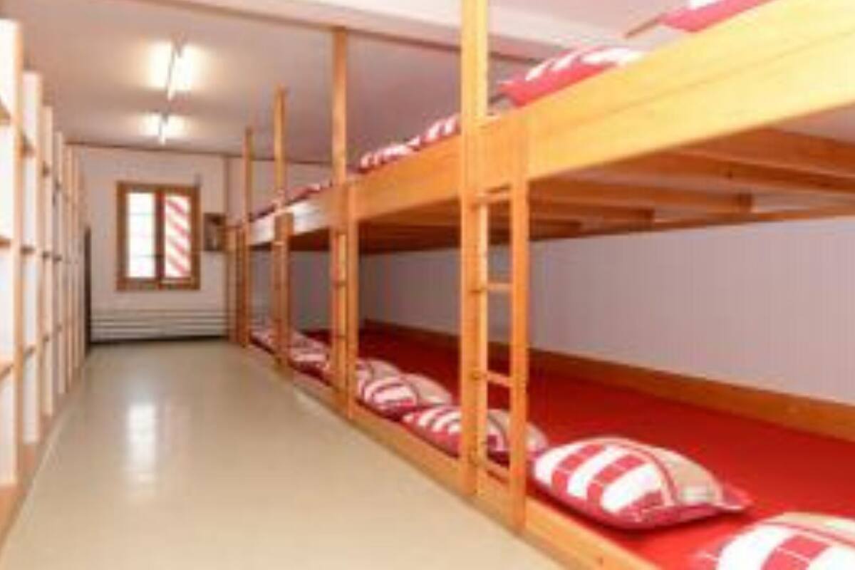 Varias literas con ropa de cama a rayas rojas y blancas en un pasillo con suelo de madera y paredes blancas.