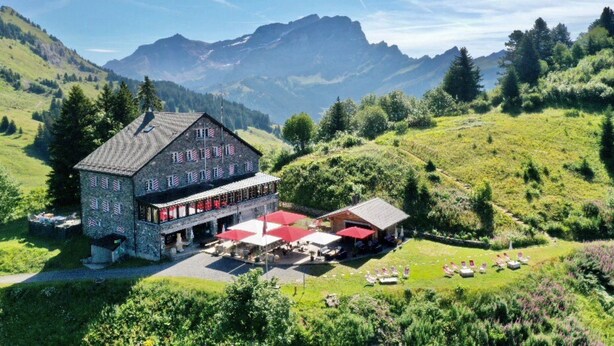 A stone building with a large terrace and outdoor seating area, surrounded by lush green hills and mountains. - La Maison de la Montagne (Ollon)