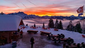 A snow-covered outdoor area with wooden benches and tables, a wooden building, and a flag. - La Maison de la Montagne (Ollon)