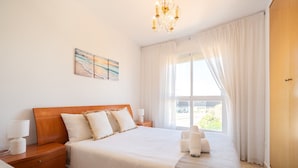 Un dormitorio con cabecera de madera, cama grande con ropa blanca, mesita de noche con lámpara, ventana con cortinas translúcidas y un cuadro en la pared.