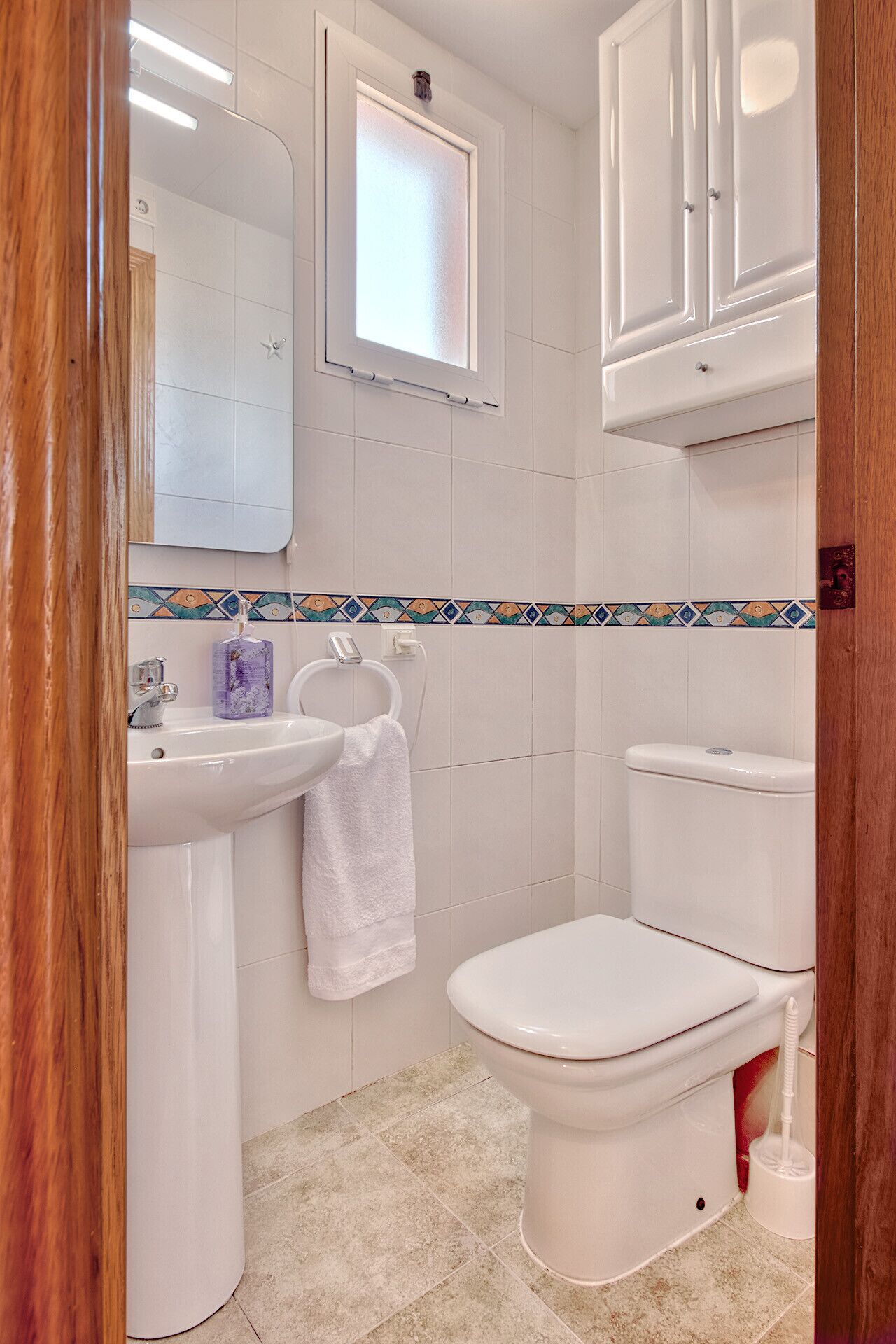 A bathroom with a white sink, a toilet, and a mirror. There is a window above the mirror, and a towel is hanging on the wall.
